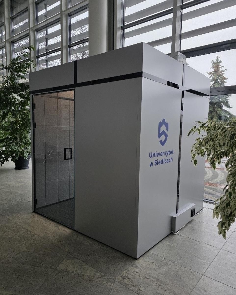 Szara kabina akustyczna w holu uczelni z widocznym logo Uniwersytet w Siedlcach na bocznej ścianie.