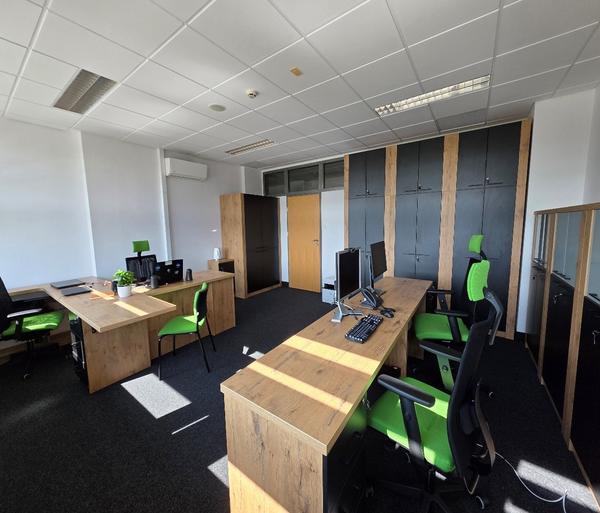 Nowe biuro Centrum Wsparcia Osób z Niepełnosprawnościami z drewnianymi biurkami, zielonymi krzesłami z kilkoma stanowiskami pracy, szafami na dokumenty i jasnym, dobrze doświetlonym wnętrzem.
